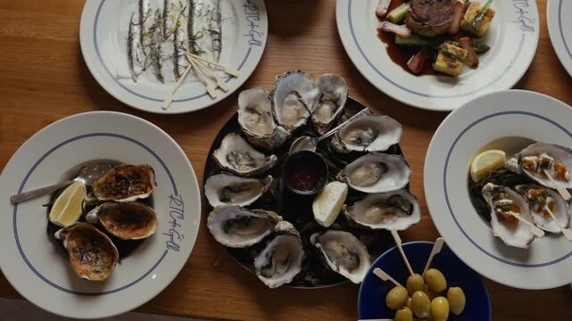 Wood table holds seafood and burger dishes. Oysters sit open, lemon wedges beside them. Plates hold grilled fish, steak, and green vegetables. No people are visible, so no emotions to describe