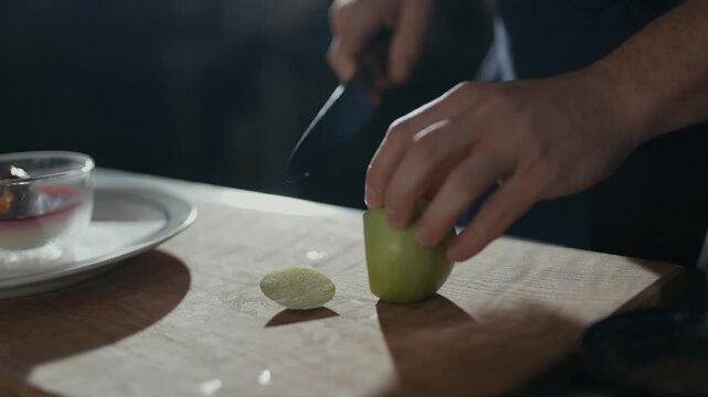 Hands slice green apple on wooden board. Light casts shadows across workspace. Dish and plate rest nearby, subtly blurred. Calm, focused action. Ideal for kitchen, cooking, or food prep scenes