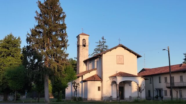 Small Italian church with bell tower in rural village, warm sunlight on traditional architecture surrounded by trees, peaceful countryside scene and cultural heritage landmark.
