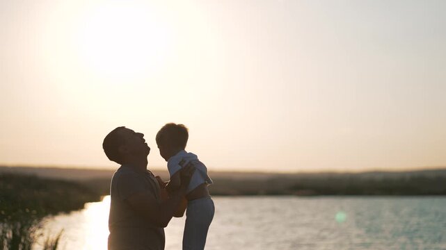 Father lifting child up in air at sunset by lake outdoors. Parent playing with son during golden hour. Family bonding time in nature. Dad holds boy high while playing together. Loving father with kid.