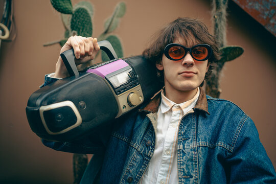 Retro fashion portrait of stylish young man with portable stereo, cassette and CD player, tinted sunglasses and denim jacket, expressing youth culture, music nostalgia and vintage lifestyle.