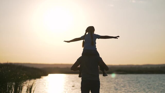 Father carrying daughter on shoulders at sunset by lake. Parent with child spreading arms wide. Dad supports girl while she pretends to fly. Family enjoys outdoor time at golden sunset hour.