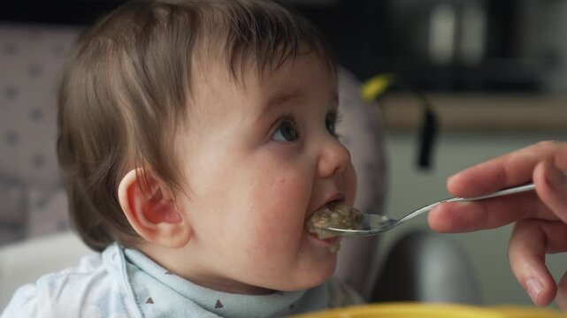 Baby eating food from spoon during feeding time. Infant enjoys nutritious meal in highchair. Parent feeds young child with spoon. Toddler eats breakfast porridge with messy face in home kitchen.