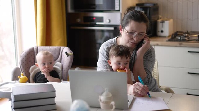 Mother multitasking with phone call while working on laptop with toddler on lap. Baby sits in highchair nearby. Mom balances remote work with childcare. Parent manages career from home kitchen.