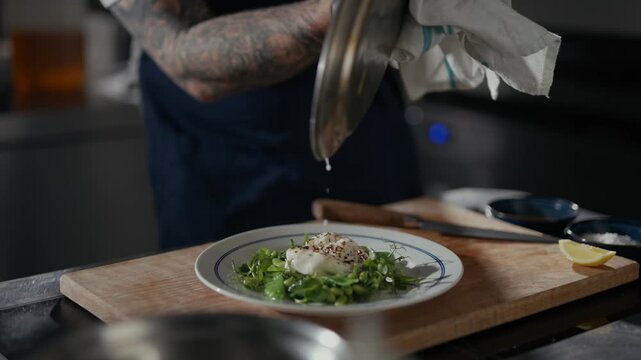 Tattooed chef drizzles finishing oil over greens. Serving dish sits on wooden board. Focus on hands and care in preparation. Ideal for food, restaurant, or cooking tutorials