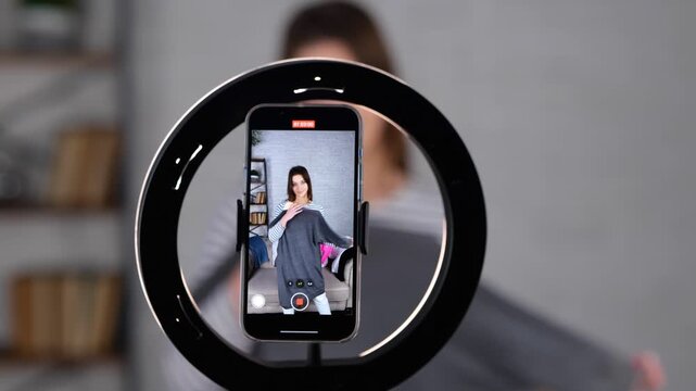 Smiling female vlogger recording video for her subscribers while holding and showing new clothes at home. Woman filming advertisement indoors. Real time video.
