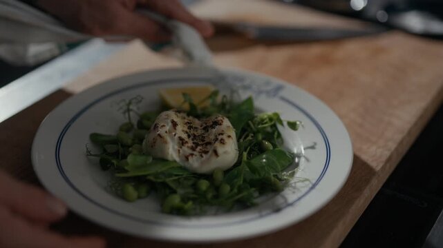 Hands hold a dish with greens and a poached egg. Lemon wedge rests beside the egg. The plate sits on a wooden board. The scene suggests a fresh, simple meal. It conveys a quiet, home-cooked atmosphere