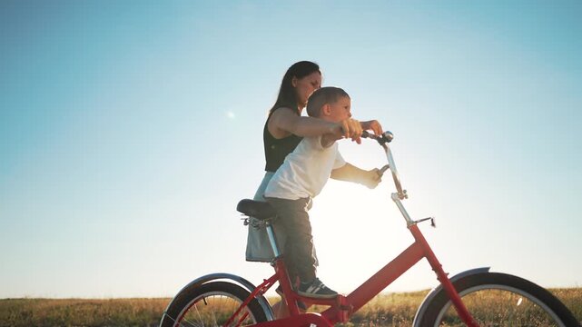 Mother teaching child to ride bicycle in sunny field. Mom helps boy learn cycling at sunset. Family bike ride outdoors. Child grips handlebar with mother guiding. Bicycle lesson in open field at sun.