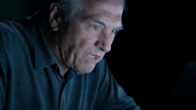 Elderly man typing on laptop in dark environment. Senior freelancer working late on computer project. Serious businessman focused on digital tasks at night time.