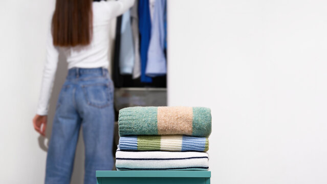Folded knitted sweaters with woman near open wardrobe on light gray background. Concept of wardrobe organization, seasonal changeover, clothing sorting, home storage and tidy routine. Copy space