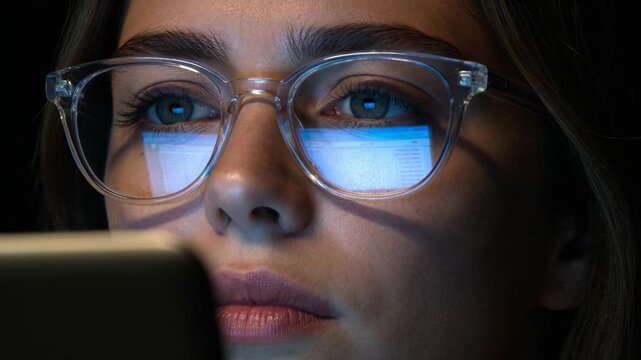 Woman wearing glasses stares at glowing screen in darkness. Close up of eyes reflecting digital light. Focus and concentration while working on computer at night.