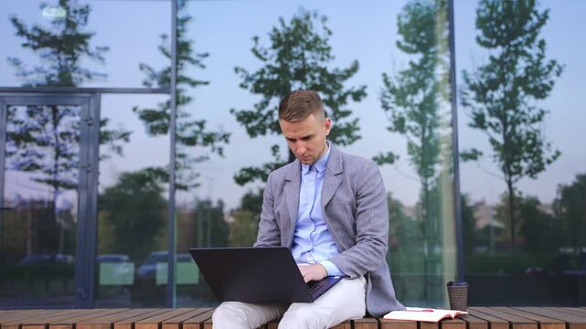 Young man sitting on the bench and typing on laptop outdoors. Real time