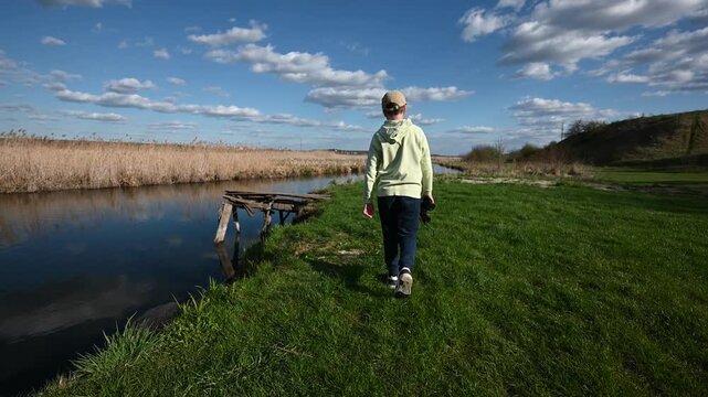 Young Boy Walks Along a Peaceful River Bank Towards a Wooden Pier, Gazing at the Expansive Landscape with Binoculars on a Sunny Day, Exploring Nature's Beauty