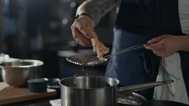 Chef removes food from hot oil with a slotted spoon. Hands show focused, precise cooking motion. Kitchen tools and pots nearby suggest active preparation. Perfect for food, culinary