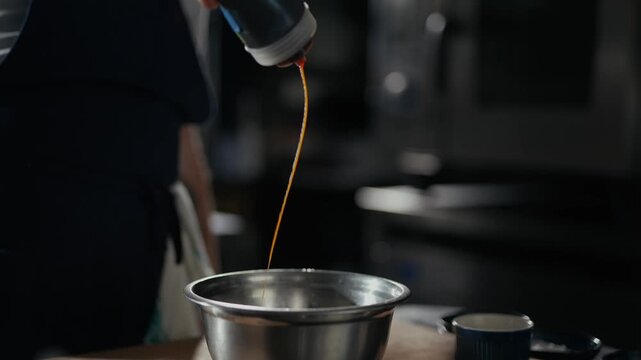 Chef pours vibrant sauce into a metal bowl with precision. Dark kitchen backdrop highlights culinary action. Professional movement suggests expert technique. Ideal for food, cooking