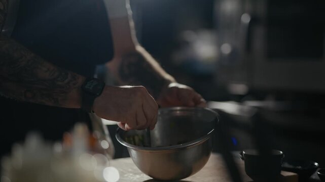 Tattooed hand stirs greens in a metal bowl. Dense shadows frame a focused, culinary motion. Kitchen tools blur softly in background light. Perfect for food prep, chef close-ups, or kitchen ambiance