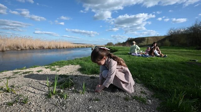 Little Girl Squatting and Playing by a Scenic Riverbank While Her Family Enjoys a Picnic on a Bright Spring Day