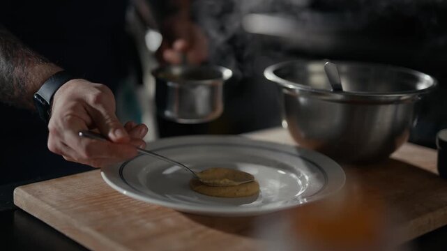 Hand scoops sauce onto a plate. Cooking scene focuses on the dish preparation. Steam rises from the kitchen background. Food presentation feels deliberate and careful