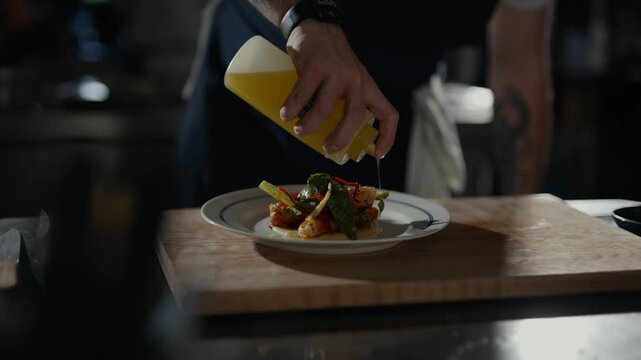 Hand pours golden liquid over plated dish. Plates sit on wooden board. Dish features greens and cooked elements. Cinematic lighting suggests fine dining. Perfect for food or culinary content