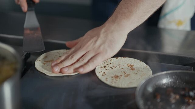 Hand flips tortilla over a hot griddle. Tortilla sizzles as it cooks on heated surface. Cook removes tortilla with spatula. Warm, steamy kitchen with food preparation focus. Ideal for food, cooking