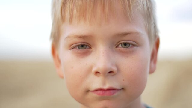 Blonde boy with green eyes looking directly at camera. Child shows different facial expressions outdoors. Young kid with blonde hair displays emotions. Boy changes expression from serious to smile.