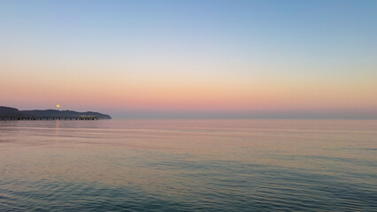 Fototapeta premium Calm sunset over the sea with a wooden pier and full moon. Peaceful twilight landscape.