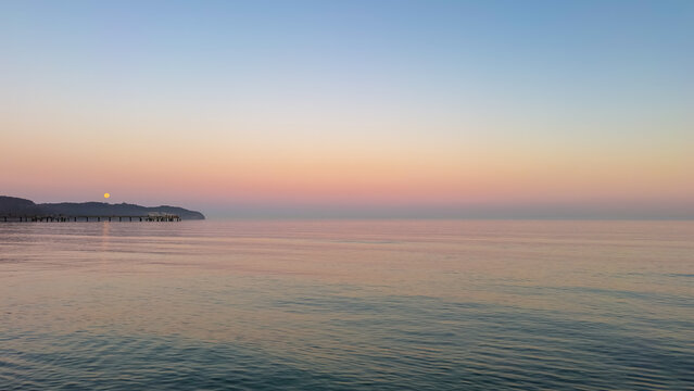 Calm sunset over the sea with a wooden pier and full moon. Peaceful twilight landscape.