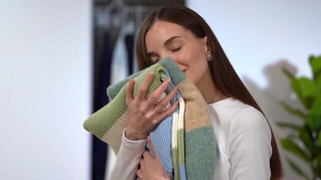 Young smiling woman pressing soft wool sweater to cheek, smelling aroma. Wardrobe, green plant on home interior background. Concept of fresh laundry routine, tactile comfort, knitwear clothing care