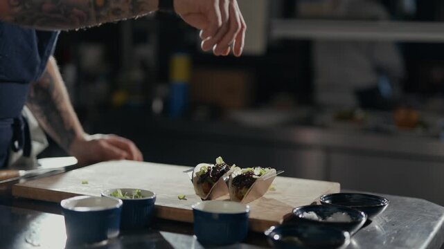 Tattooed hand sprinkles fresh greens over tacos. Cook's focused action adds final garnish. Professional kitchen setting, warm lighting. Perfect for food prep, culinary branding