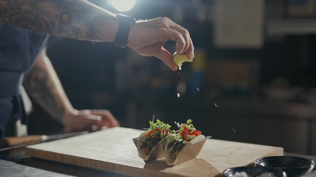 Hands season tacos with care. Tattooed arms move with precision. Food rests on a wooden board. Light shines from a single source. Taco preparation in a dim kitchen. Great for food, cooking
