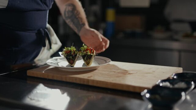 Chef's tattooed hands place tacos on a plate. Hands show care in final plating. Bright kitchen lights highlight textures. Perfect for food blogs or restaurant ads