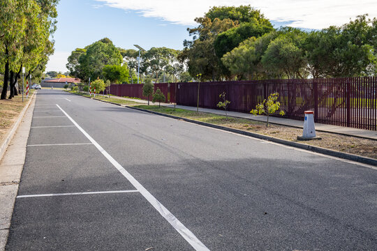 A quiet suburban street with parallel parking bays marked in  lines along&nbsp;road adjacent to a school campus perimeter in Melbourne, Australia.&nbsp;Paling security fence borders the school boundary