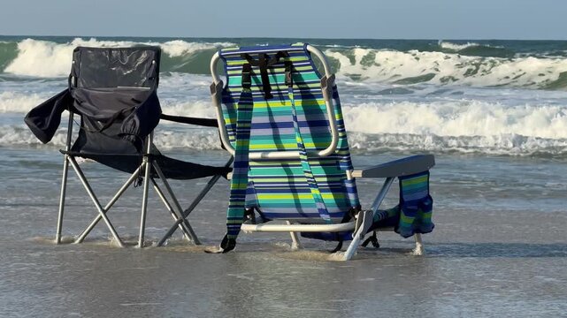Foldable beach chairs sit in shallow surf as waves roll toward the shore. Quiet seaside composition suggests relaxation, vacation, and peaceful coastal living.
