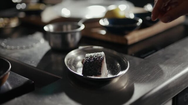 Hand sprinkles salt over delicate food item. Metal surface reflects kitchen tools softly. Focus sharpens on culinary precision. Professional kitchen setting suggests high-end preparation