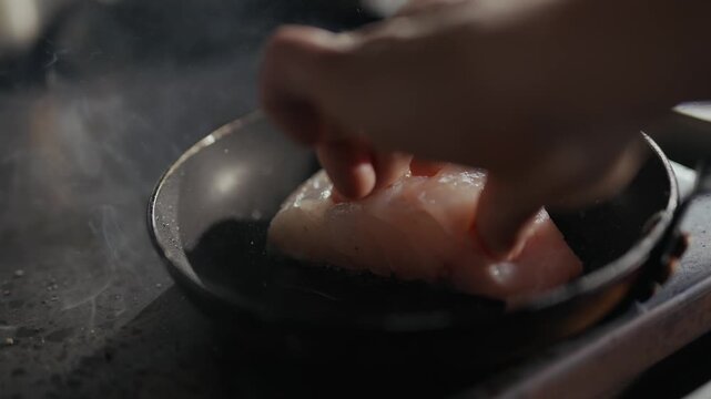 Hand presses fish into hot pan. Steam rises from the pan. Fingers gently handle the fish. Cooking process shown closely. Natural, simple meal prep moment