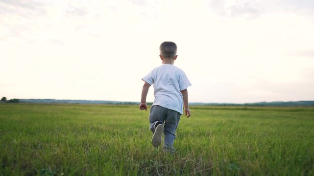 Little boy running through green grass field. Child runs freely across open meadow at sunset. Kid enjoys outdoor freedom on summer evening. Boy sprints through lush grassland under cloudy sky.