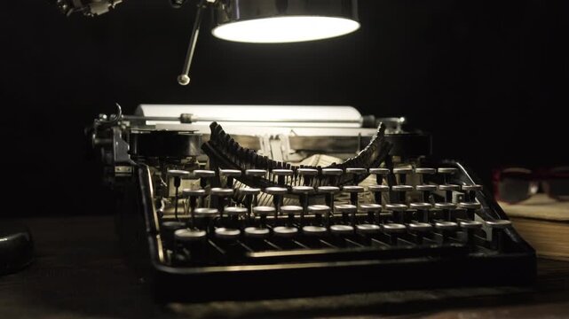 Old vintage typewriter lit by table lamp on desk in dark office rotating on black background close-up