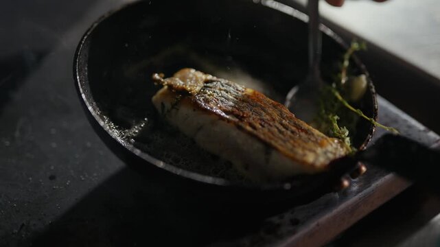 Chef adds herbs to seared fish in cast iron skillet. Sizzle of oil and garlic fills the air. Golden crust forms over delicate fillet. Perfectly cooked fish hints at fresh, savory meal