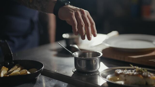 Chef sprinkles herbs over a dish with care. Hand hovers above food, ready to finish cooking. Professional kitchen setting, dark tones, focused lighting. Perfect for culinary, food prep