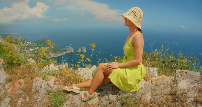 A woman sits on a hill overlooking blue sea and Faraglioni sea stacks on Capri Italy