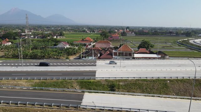 Aerial view of a modern highway passing through rural landscapes with Mount Merapi in the distance and a car driving on the road in Klaten, Central Java, Indonesia.