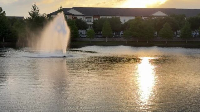 A tall water fountain rises from a quiet neighborhood lake with warm sunset reflections. Trees and apartment buildings create a peaceful suburban evening landscape.