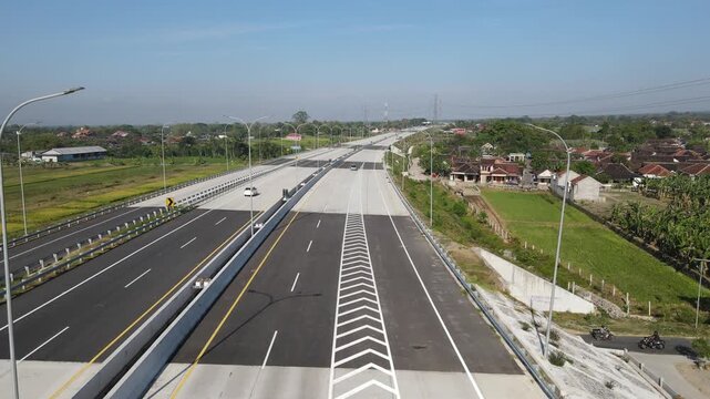 Aerial view of a modern multi-lane highway with cars driving past green fields and residential houses under a clear blue sky in Klaten, Central Java, Indonesia.