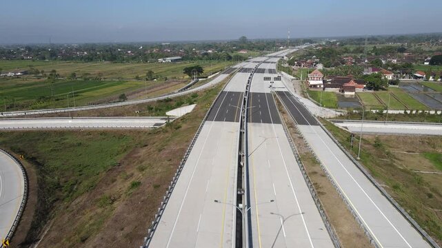 Aerial view of a modern highway interchange with multi-lane roads passing through rural landscapes and green fields under a clear sky in Klaten, Central Java, Indonesia.