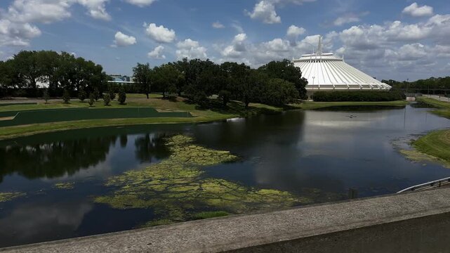 The white Space Mountain structure stands beside a reflective lagoon under a bright cloudy sky. Trees and pathways surround the iconic theme park landscape.