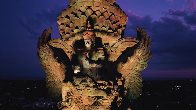 Aerial view of the illuminated Garuda Wisnu Kencana statue at night with city lights and a twilight sky in Kecamatan Kuta Selatan, Bali, Indonesia.