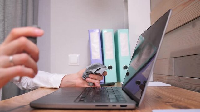 Person's hands spraying cleaning liquid on a soft cloth while preparing to wipe a laptop screen on a wooden desk in an office environment, promoting hygiene and cleanliness for electronic devices