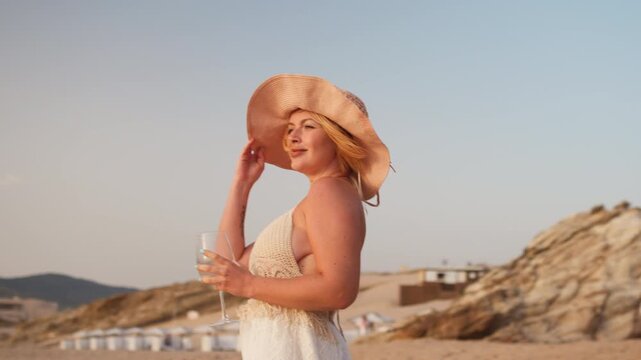 Smiling young woman in sunhat and summer dress drinking wine at sandy beach enjoying holiday leisure and breeze at sunset