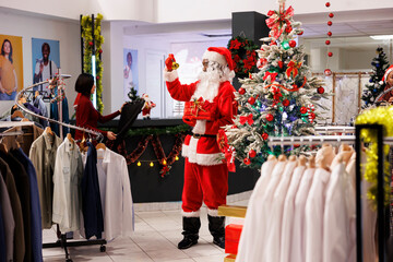 Fototapeta premium African american man dressed as Santa Claus in festive decorated shopping mall clothing store. Cheerful employee jingling bells and holding red package in fashion boutique shop.