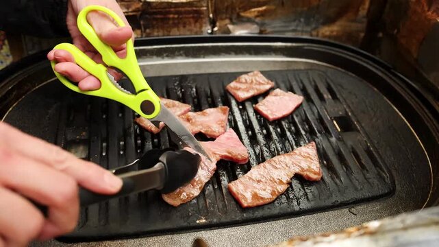 Chef using lime green kitchen scissors and tongs to cut premium wagyu beef slices on indoor electric grill.